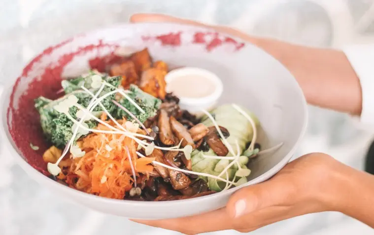 A colorful and healthy vegan bowl featuring fresh vegetables, sautéed mushrooms, cabbage, shredded carrots, kale, microgreens, and a side of dressing, held in human hands.